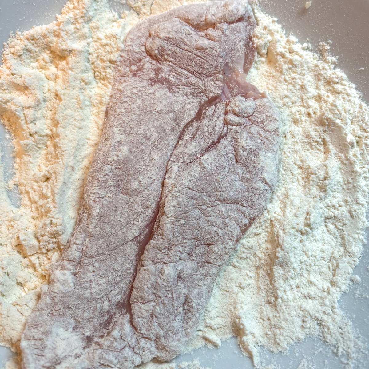 A raw chicken cutlet being coated in plain flour in a shallow dish, ready for pan-frying.