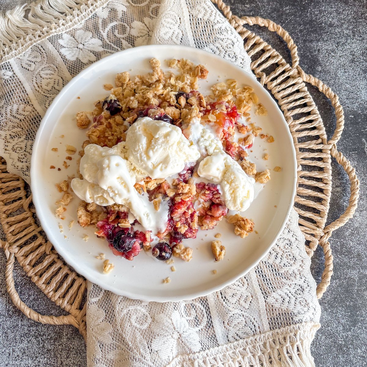 Plum and blueberry crumble with ice cream and crisp oats on a woven placemat