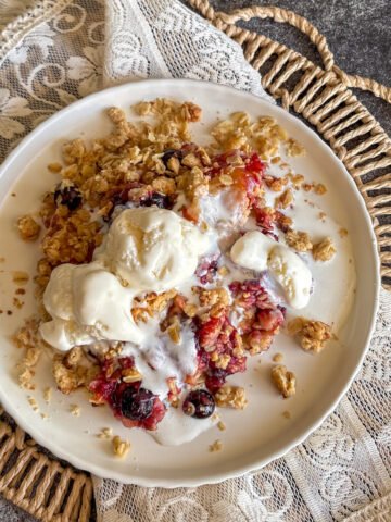 Plum and blueberry crumble with melting ice cream and golden oat topping on a white plate