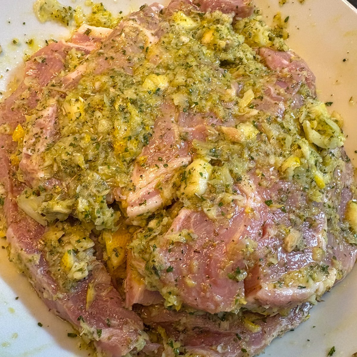 Raw pork gigot chops coated in a blitzed lemon and garlic herb marinade in a white bowl, showing the green-flecked paste rubbed into every piece before marinating.