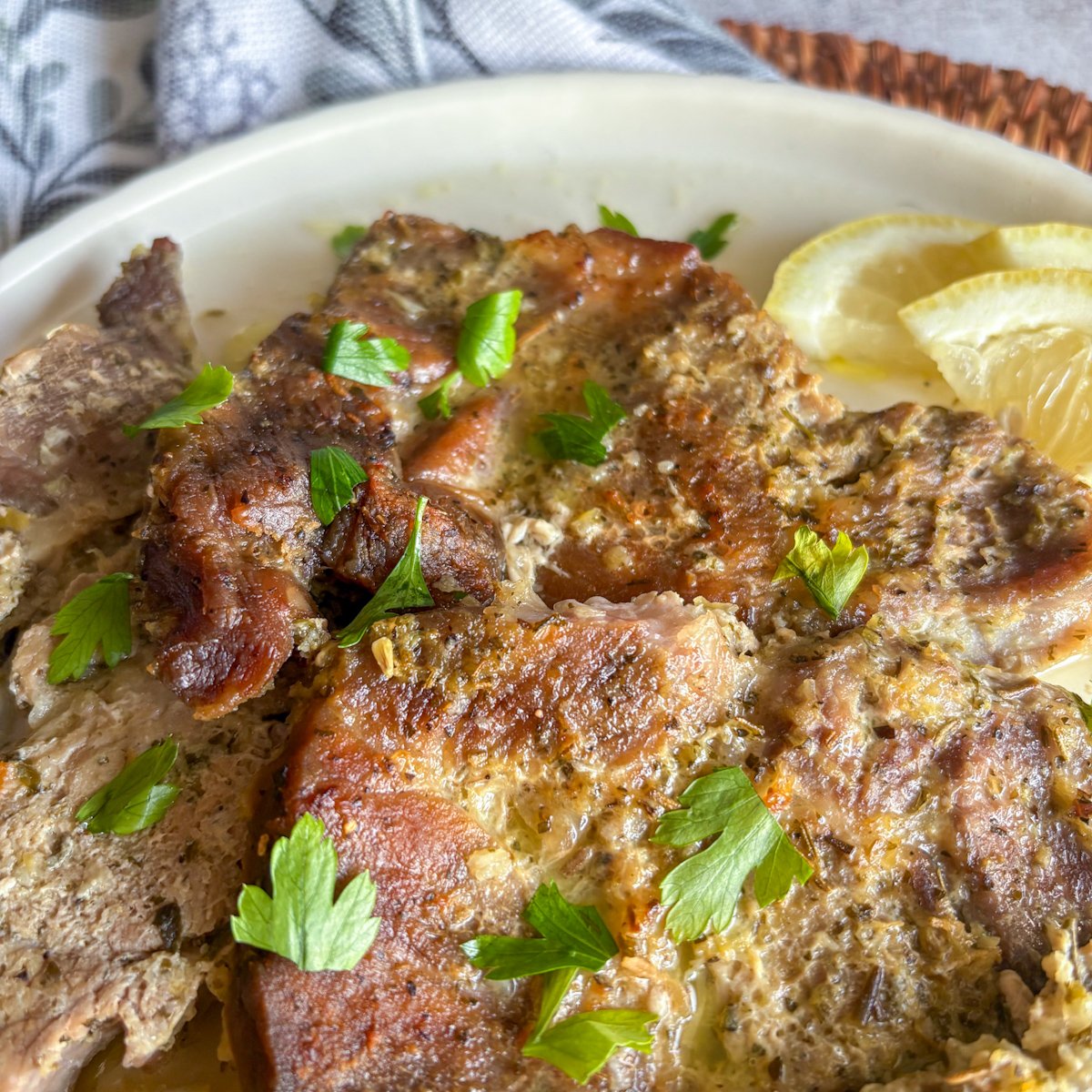 Close-up of lemon garlic pork gigot chops with browned tops and visible herb marinade, garnished with fresh parsley and two lemon slices on a cream plate.