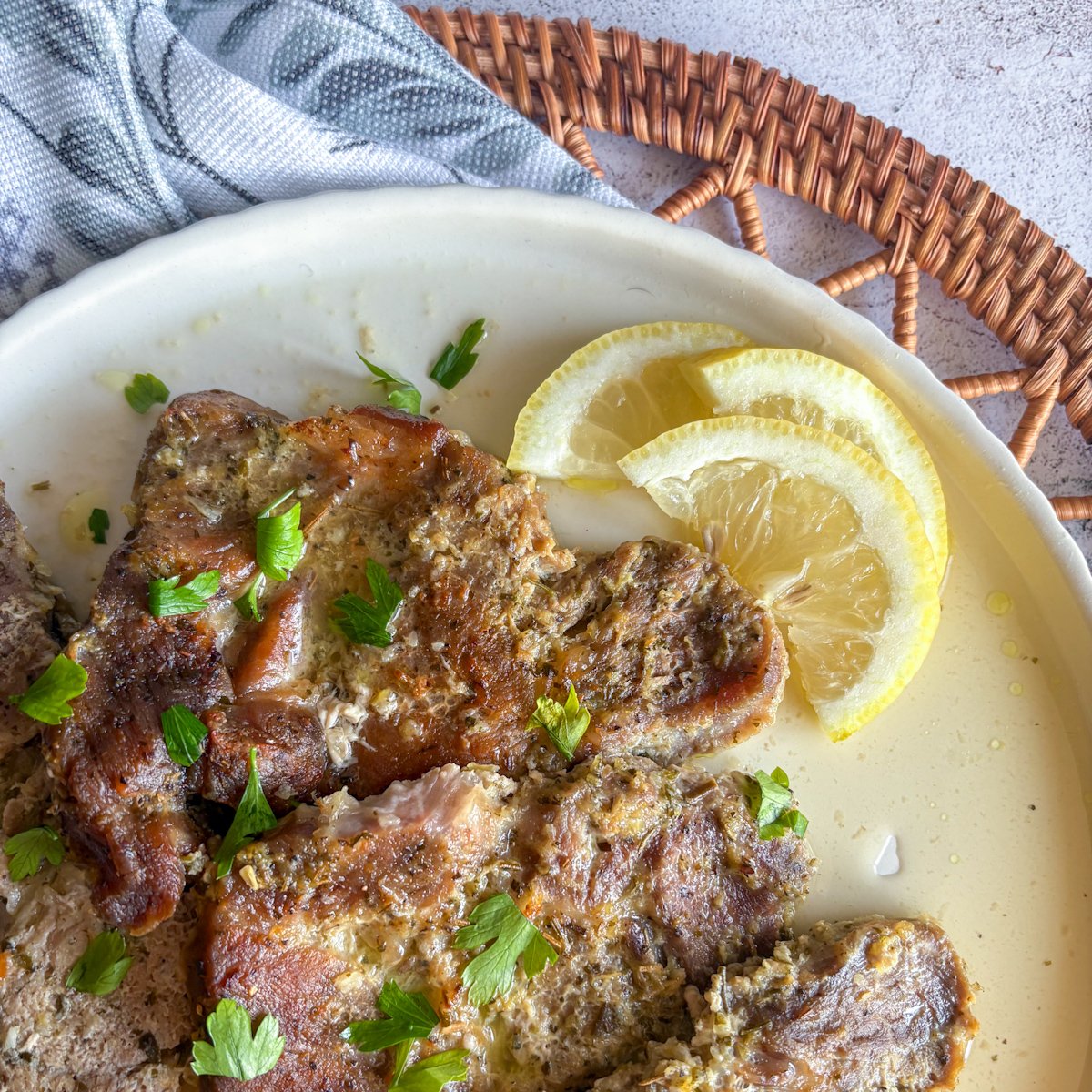 Overhead close-up of tender pork gigot chops glossy with lemony pan juices, topped with parsley and served with lemon slices on a rustic plate.