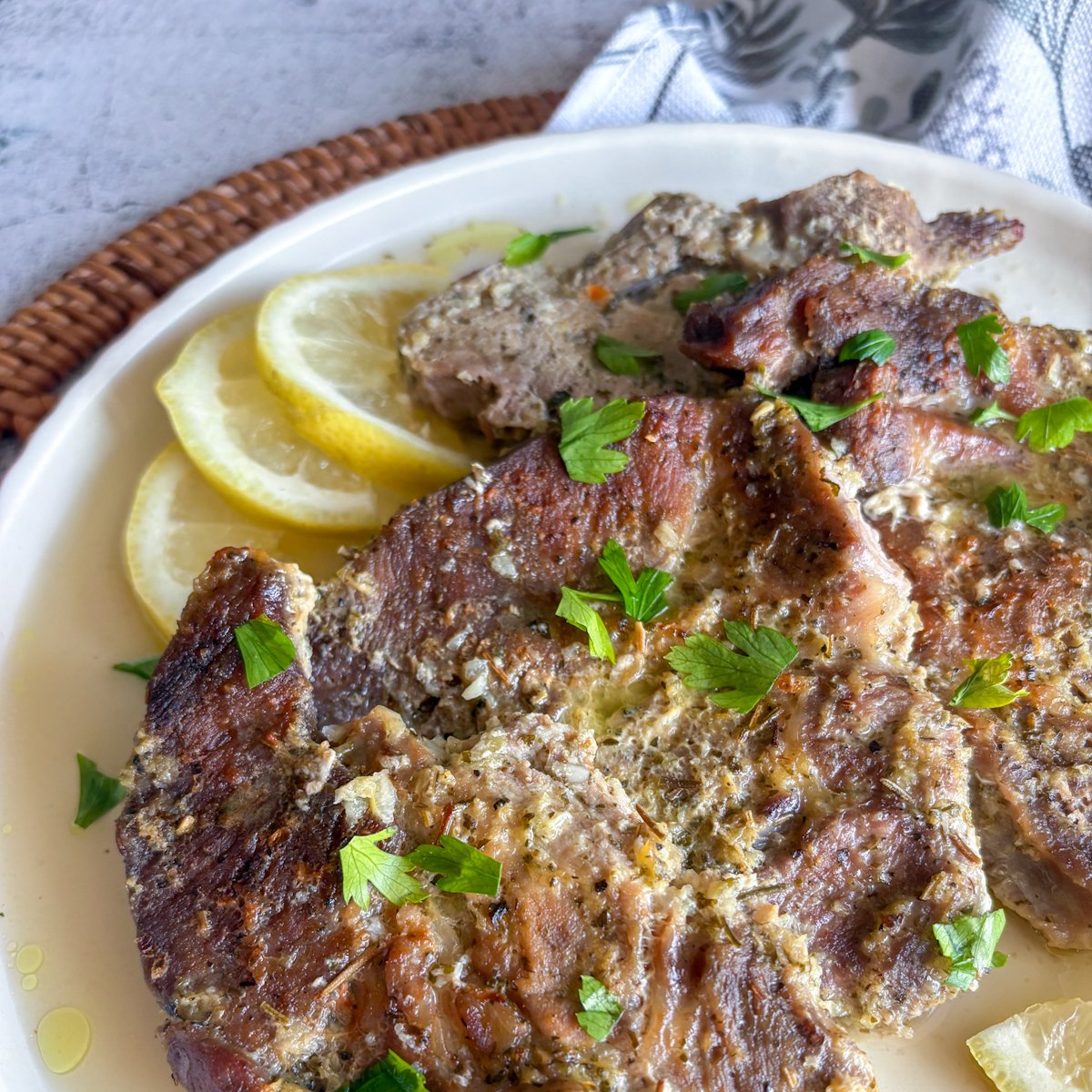 Close-up of slow braised pork gigot chops with caramelised browned surfaces and herb-flecked marinade, garnished with parsley beside lemon slices.