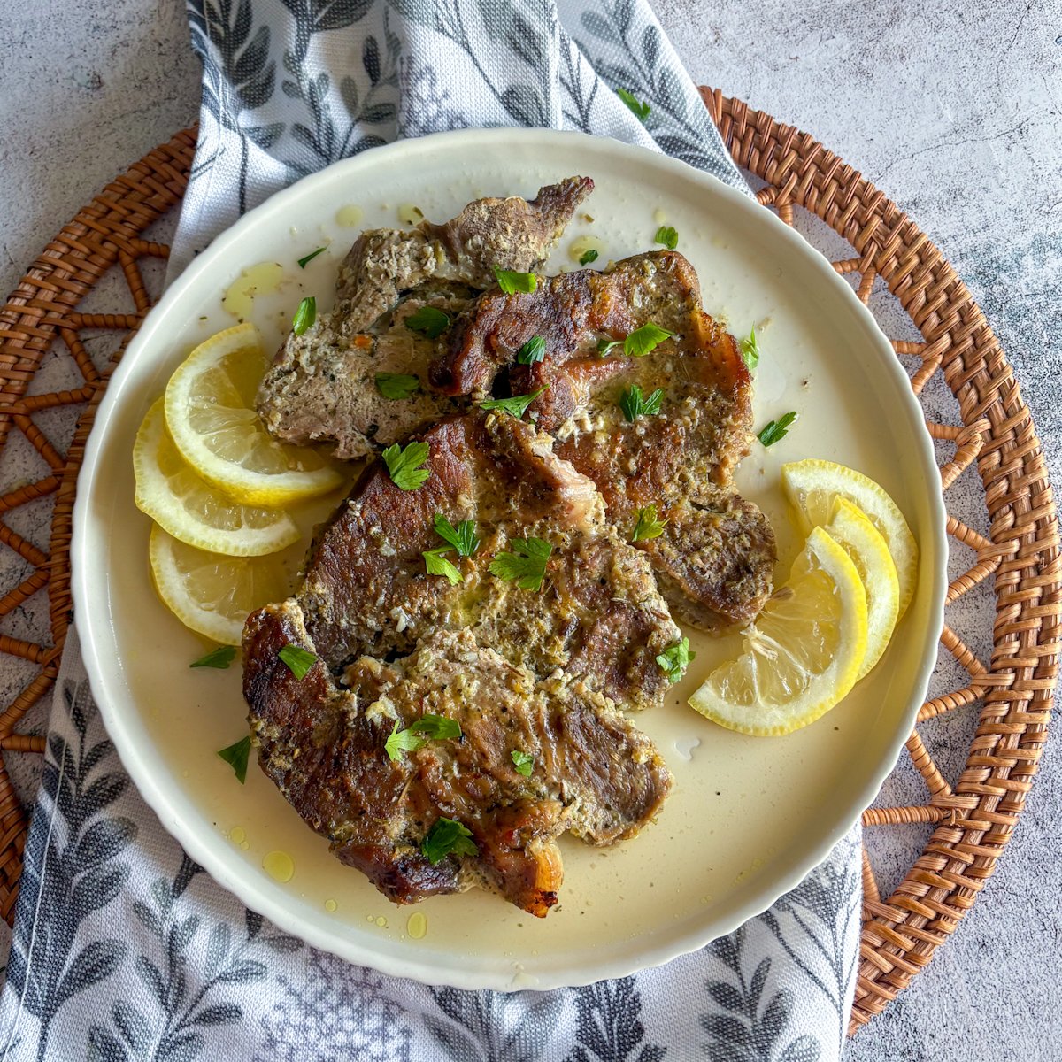 Oven braised pork gigot chops with golden browned edges and lemon garlic marinade, garnished with parsley and lemon wedges on a pale ceramic plate over a botanical cloth.