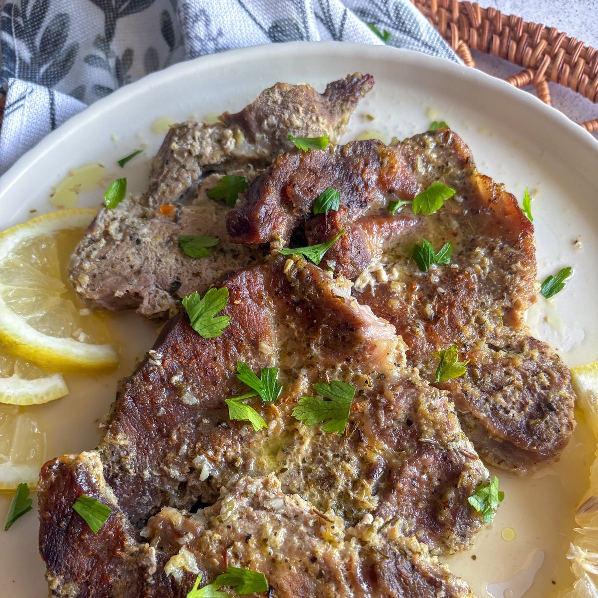 Close-up of tender pork gigot chops showing crisp browned tops and herby marinade, topped with fresh parsley beside a lemon slice on a cream plate.