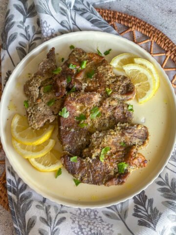Tender oven braised pork gigot chops plated on a cream scalloped dish, scattered with fresh parsley and fanned lemon slices, set on a leaf-print napkin and woven rattan mat.
