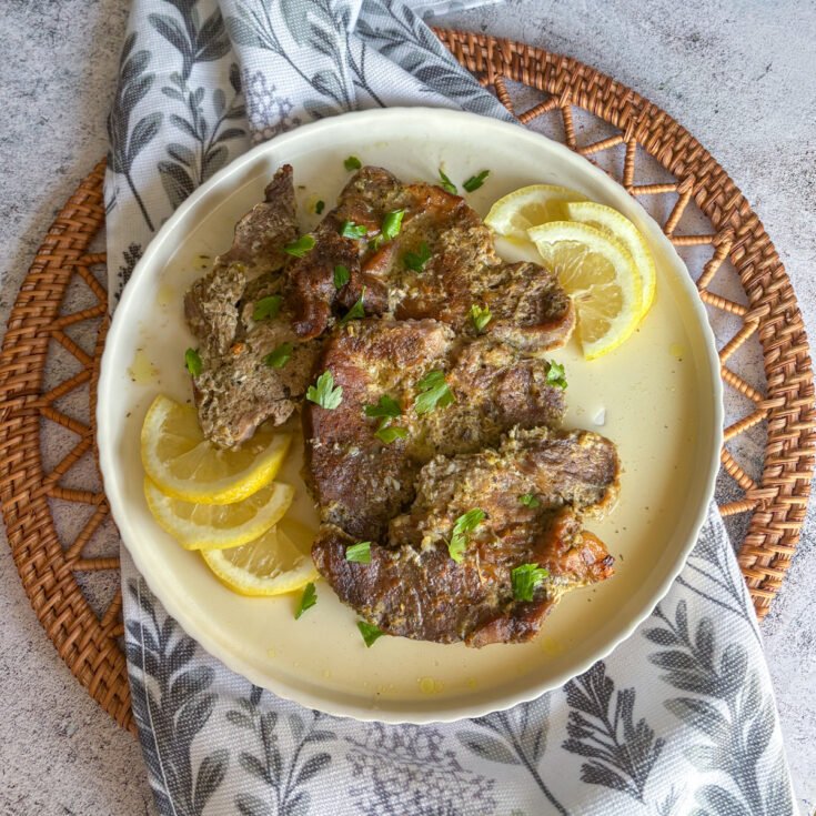 Tender oven braised pork gigot chops plated on a cream scalloped dish, scattered with fresh parsley and fanned lemon slices, set on a leaf-print napkin and woven rattan mat.
