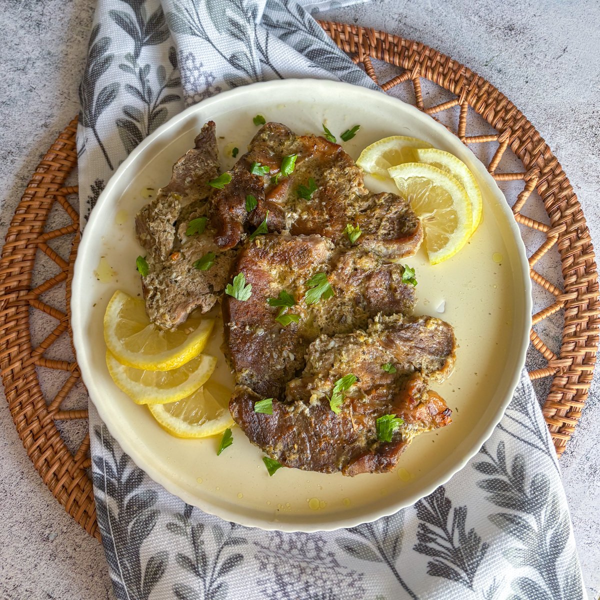 Tender oven braised pork gigot chops plated on a cream scalloped dish, scattered with fresh parsley and fanned lemon slices, set on a leaf-print napkin and woven rattan mat.