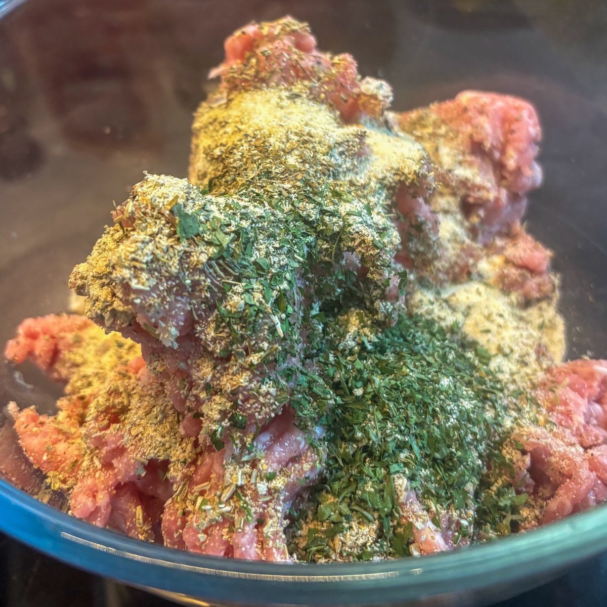 Seasoned pork mince in a glass bowl topped with dried herbs, garlic granules and onion granules before mixing.