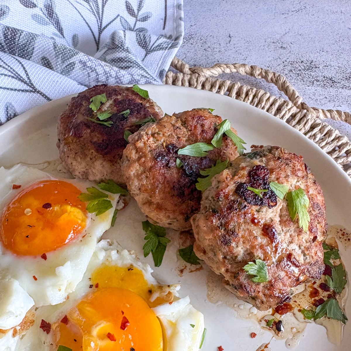 A stack of golden pork breakfast patties with fried eggs, fresh parsley and chilli flakes on a white plate.