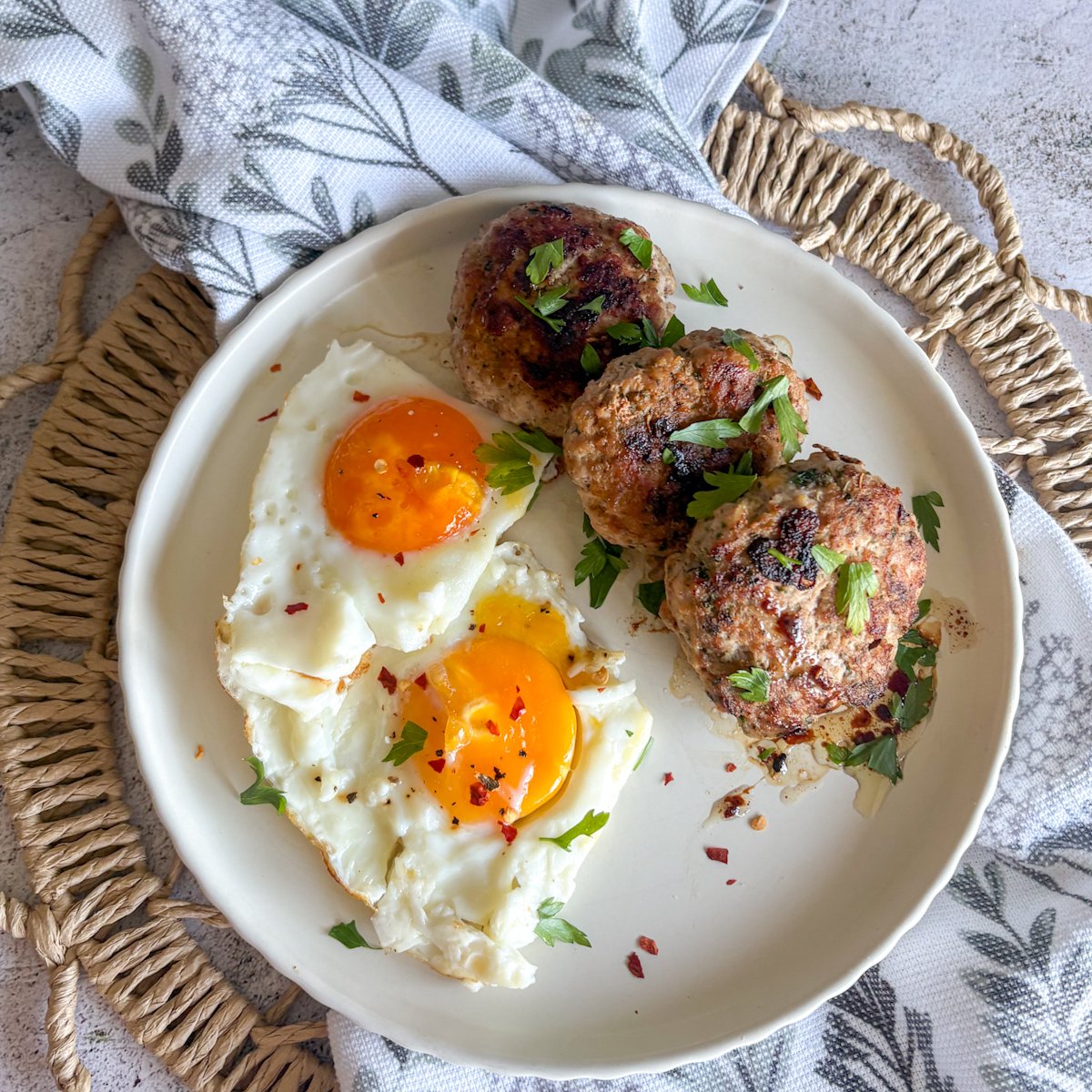 Overhead close-up of herb-flecked pork mince patties beside two fried eggs, garnished with parsley and chilli flakes.