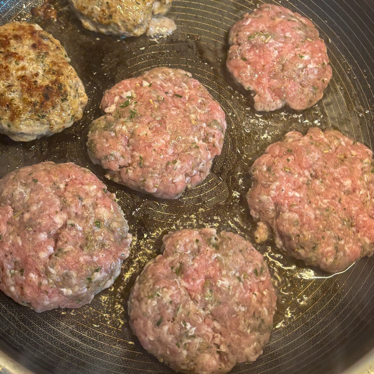 Raw pork mince patties frying in a skillet, some browned and some still pink, in a little hot oil.