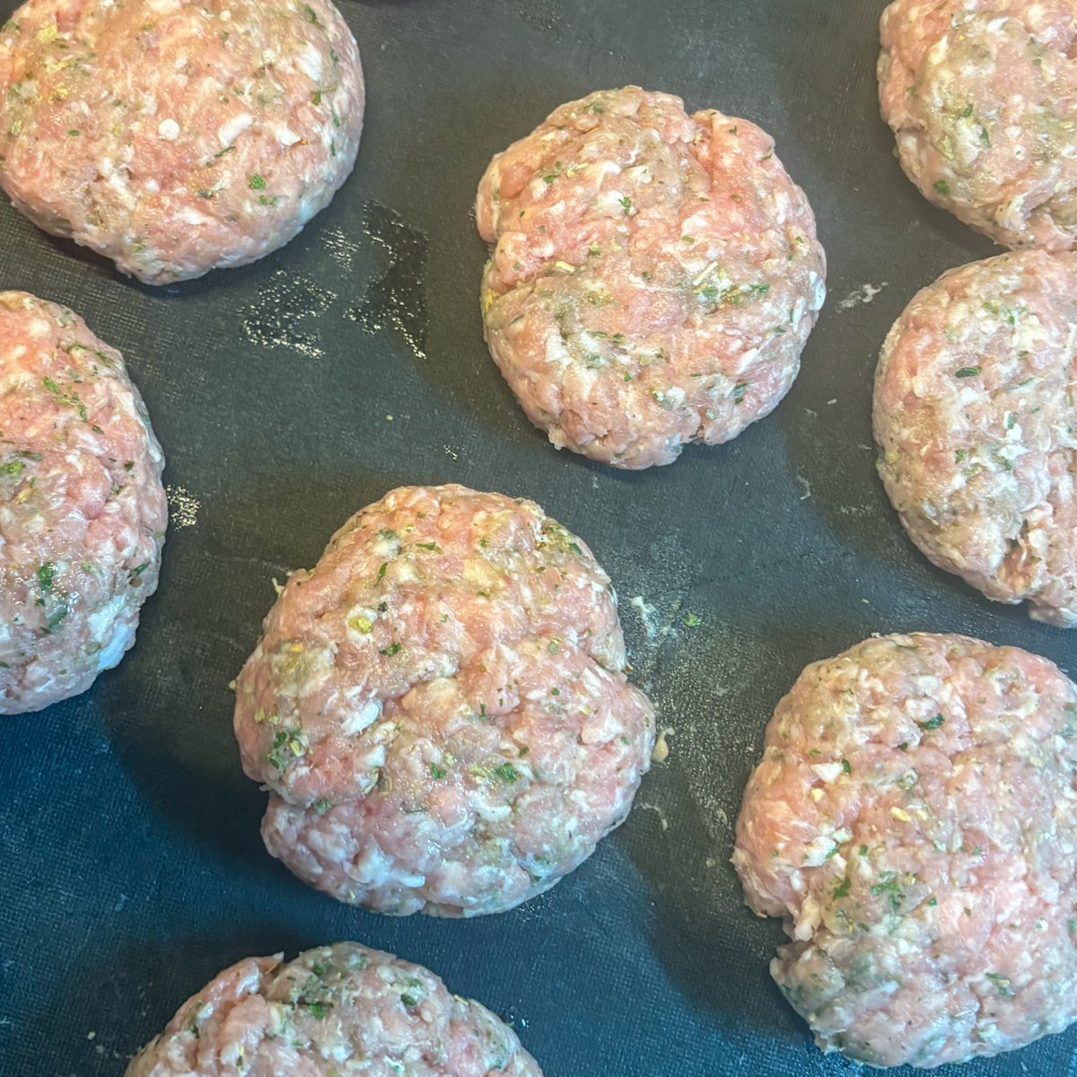 Shaped raw pork mince patties resting on a dark board before frying, flecked with dried herbs.