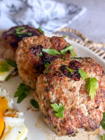 Close-up of three juicy pork mince patties topped with parsley, with runny fried eggs to the side on a white plate.