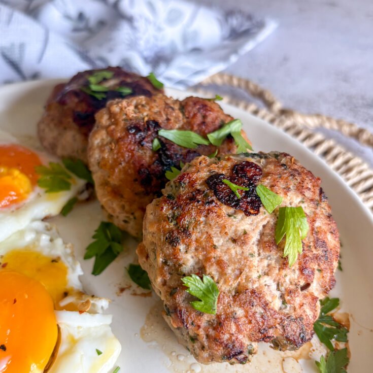 Close-up of three juicy pork mince patties topped with parsley, with runny fried eggs to the side on a white plate.