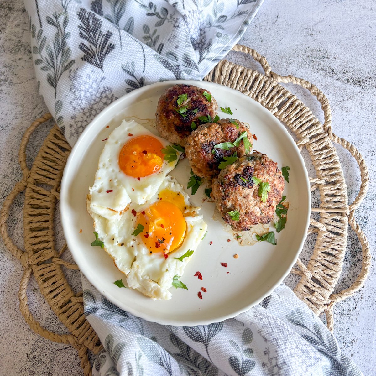 ork mince patties served on a white plate with two sunny-side-up eggs, garnished with fresh parsley and chilli flakes, on a woven mat.