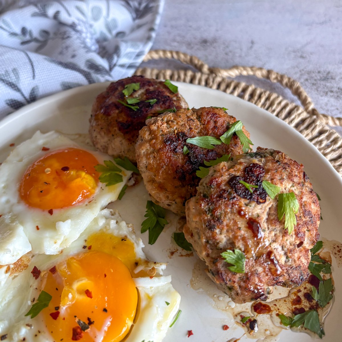Three herby pork mince patties with two sunny-side-up eggs on a white plate, sprinkled with parsley and red chilli flakes.