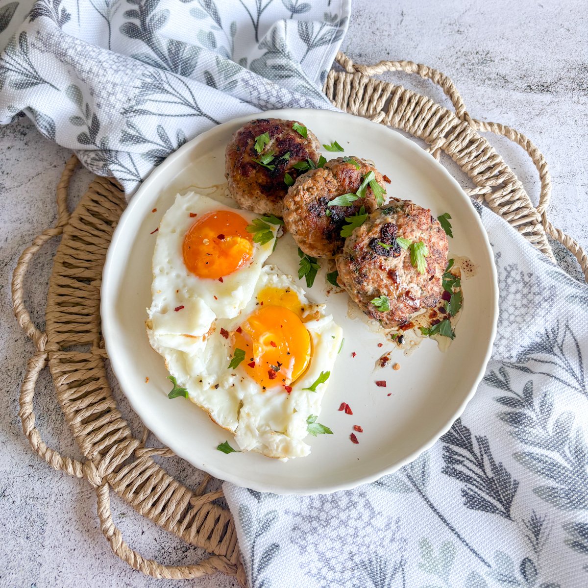 Overhead view of pork mince patties and two fried eggs on a white plate, garnished with parsley, on a floral cloth and woven mat.