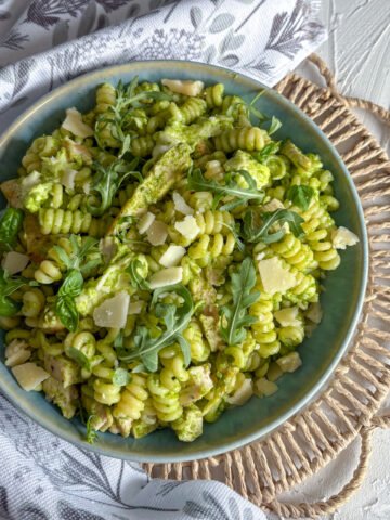 Overhead view of rocket pesto pasta salad with chicken in a teal bowl on a woven placemat, garnished with fresh rocket and basil leaves