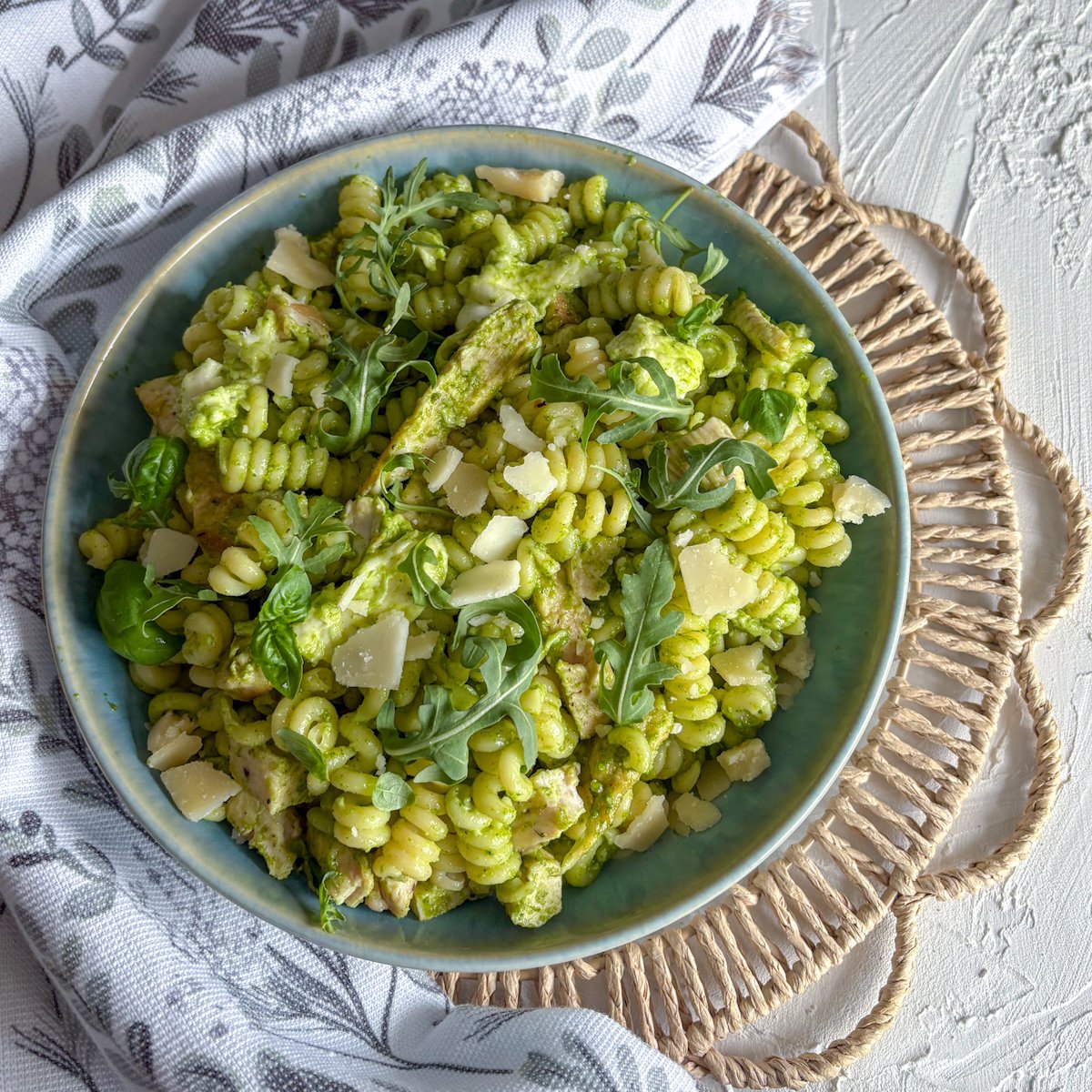 Overhead view of rocket pesto pasta salad with chicken in a teal bowl on a woven placemat, garnished with fresh rocket and basil leaves