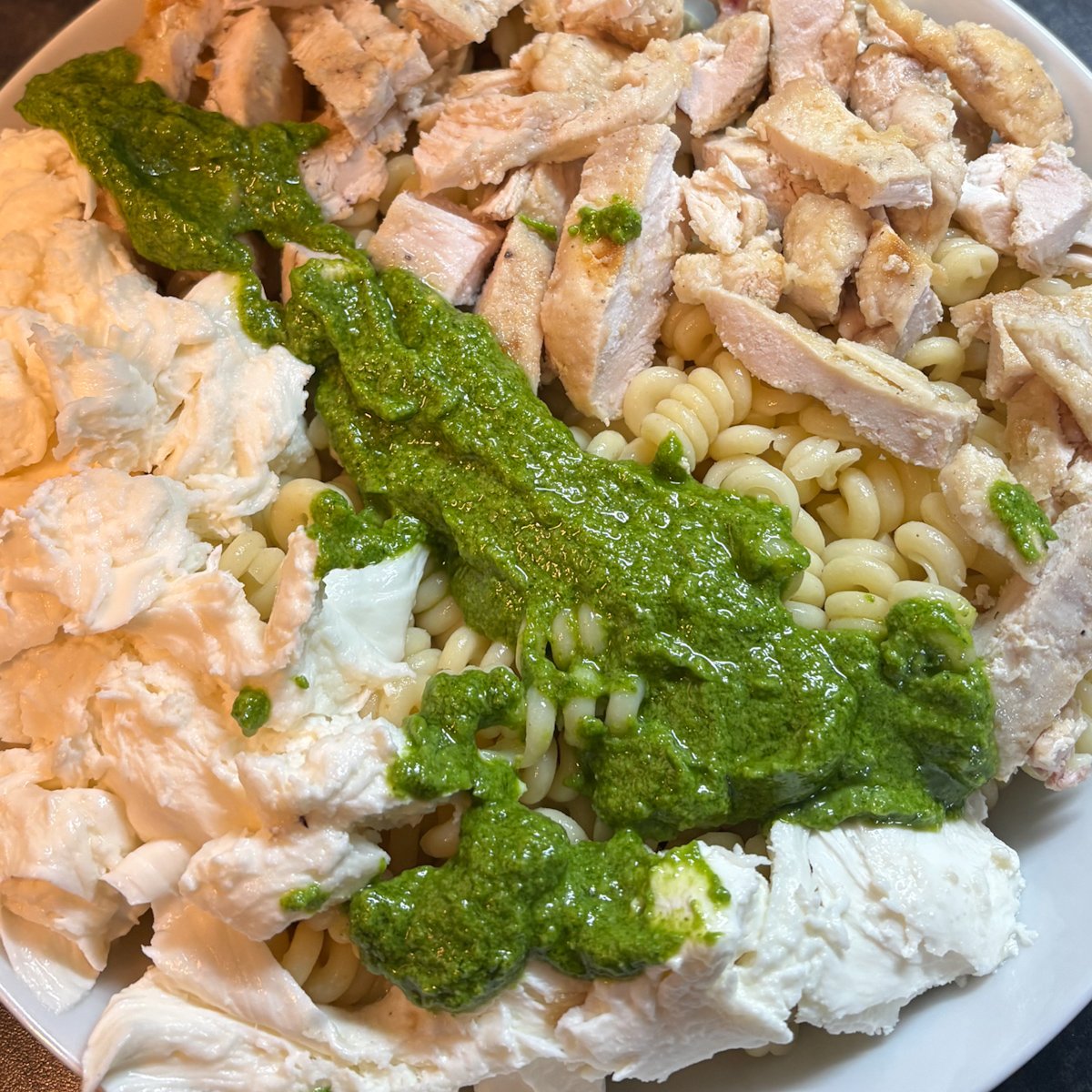 Rocket pesto pasta salad being assembled in a white bowl, with cold spiral pasta, sliced cooked chicken, torn mozzarella and green pesto poured over before mixing