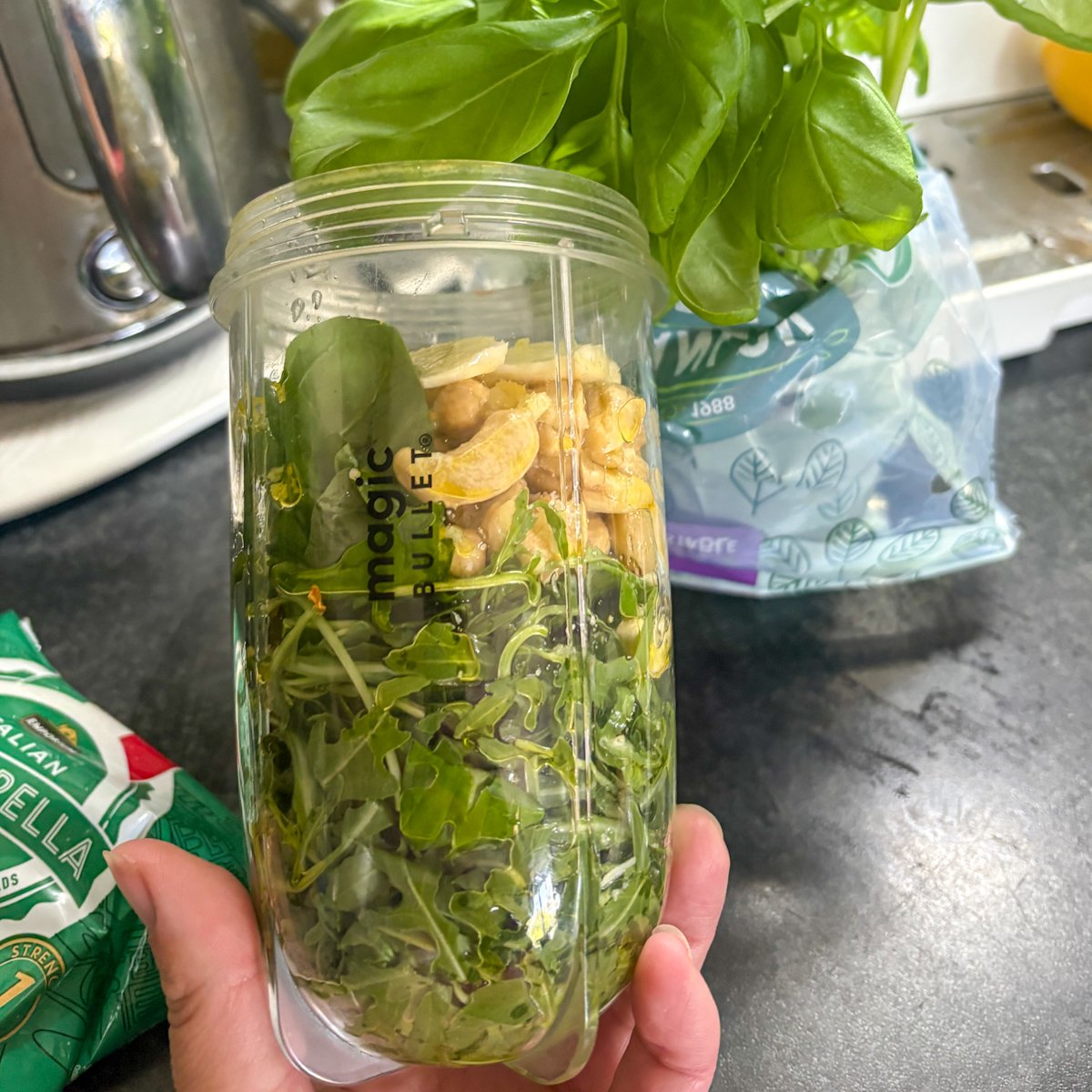 Ingredients for rocket pesto in a blender cup, with fresh rocket leaves, basil, whole cashew nuts and peeled garlic cloves waiting to be blended