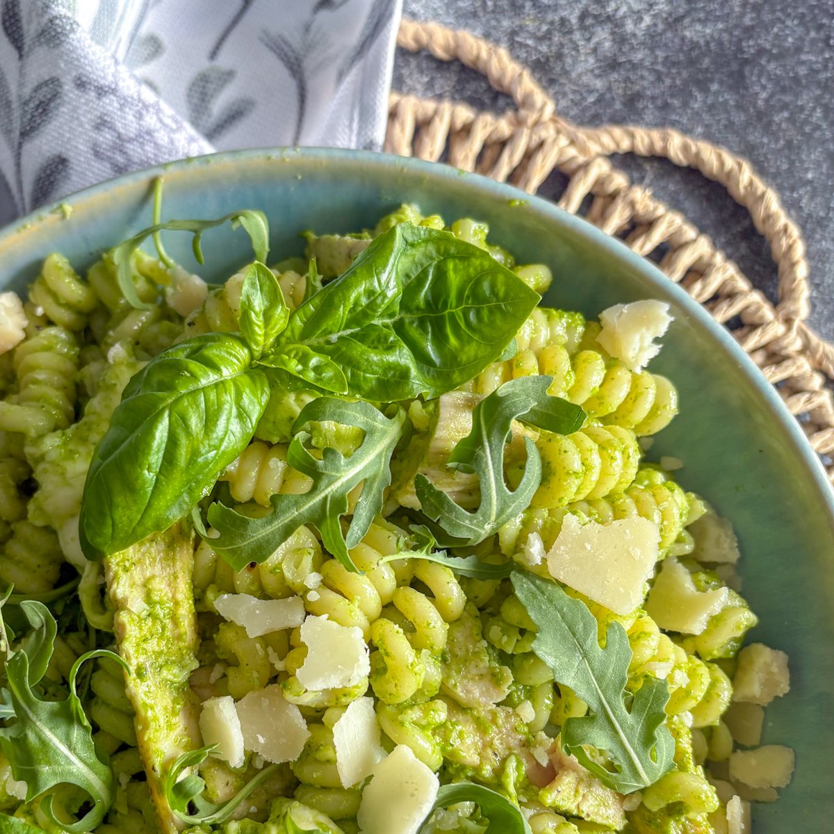 Close-up of rocket pesto pasta salad in a teal bowl, with spiral pasta coated in creamy green pesto, strips of chicken, parmesan shards and a large basil sprig on top
