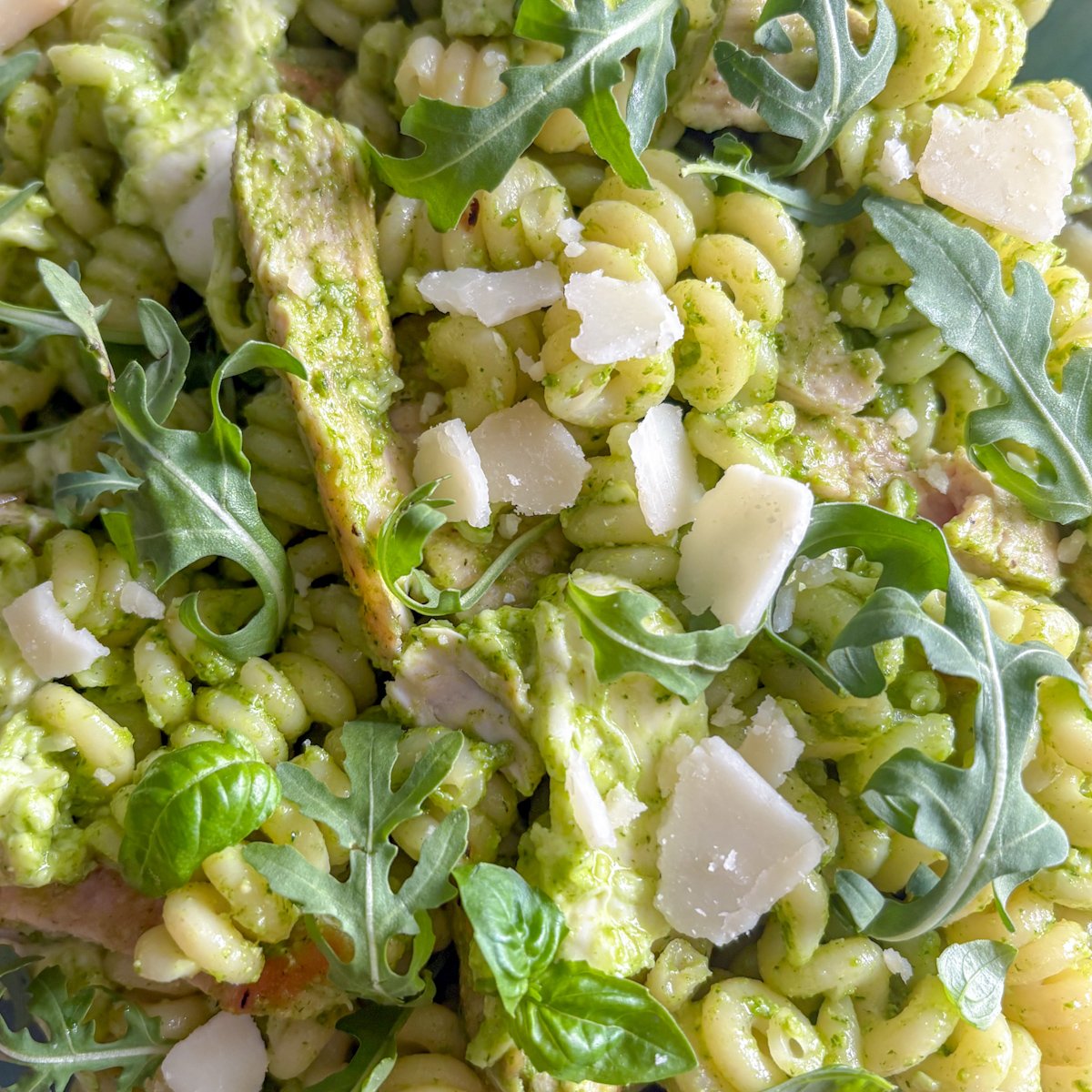 Close crop of rocket pesto pasta salad showing a sliced chicken strip coated in green pesto, surrounded by spiral pasta, peppery rocket leaves and parmesan