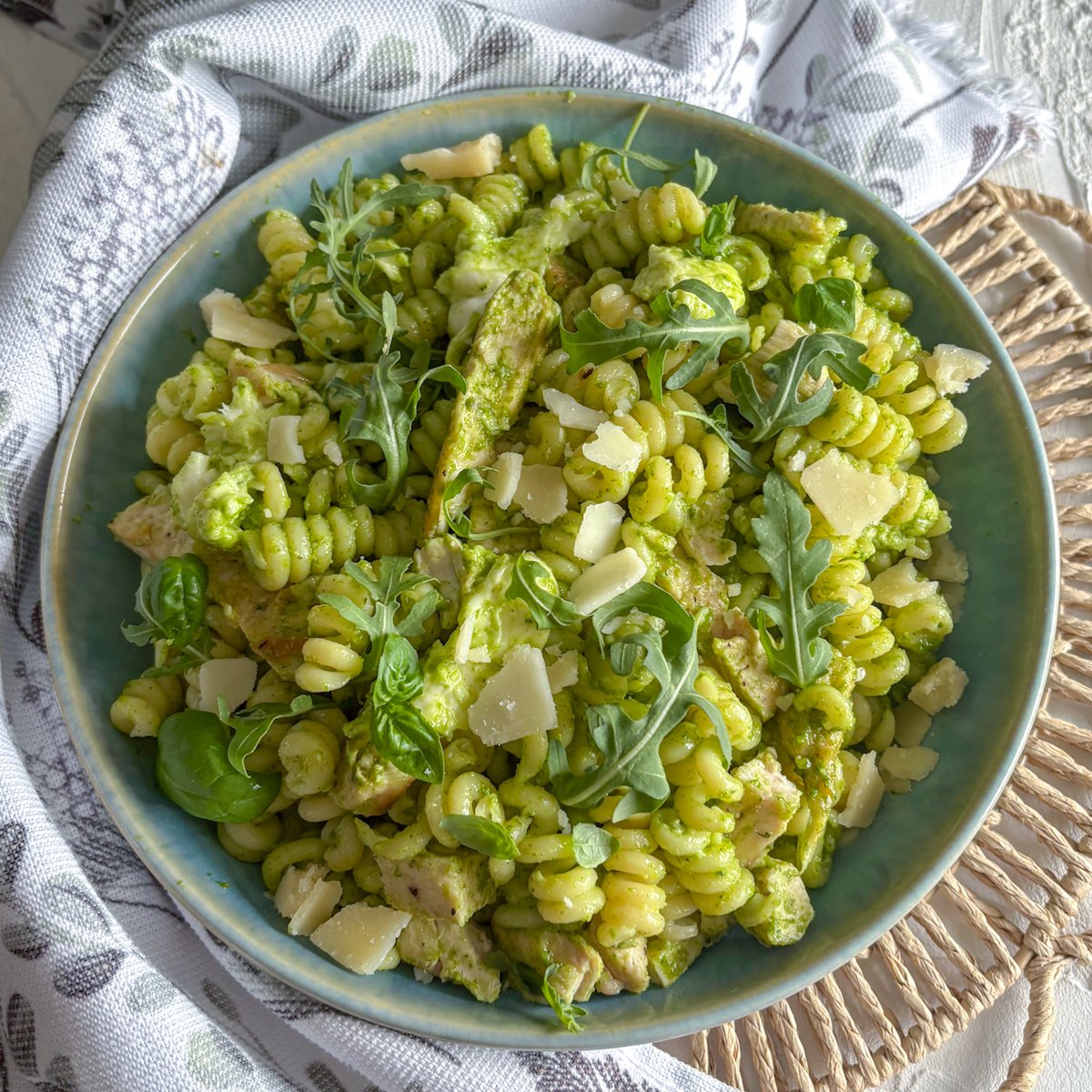 Rocket pesto pasta salad served in a blue-green bowl on a linen tea towel, with chicken, mozzarella, rocket and shaved parmesan mixed through the pasta
