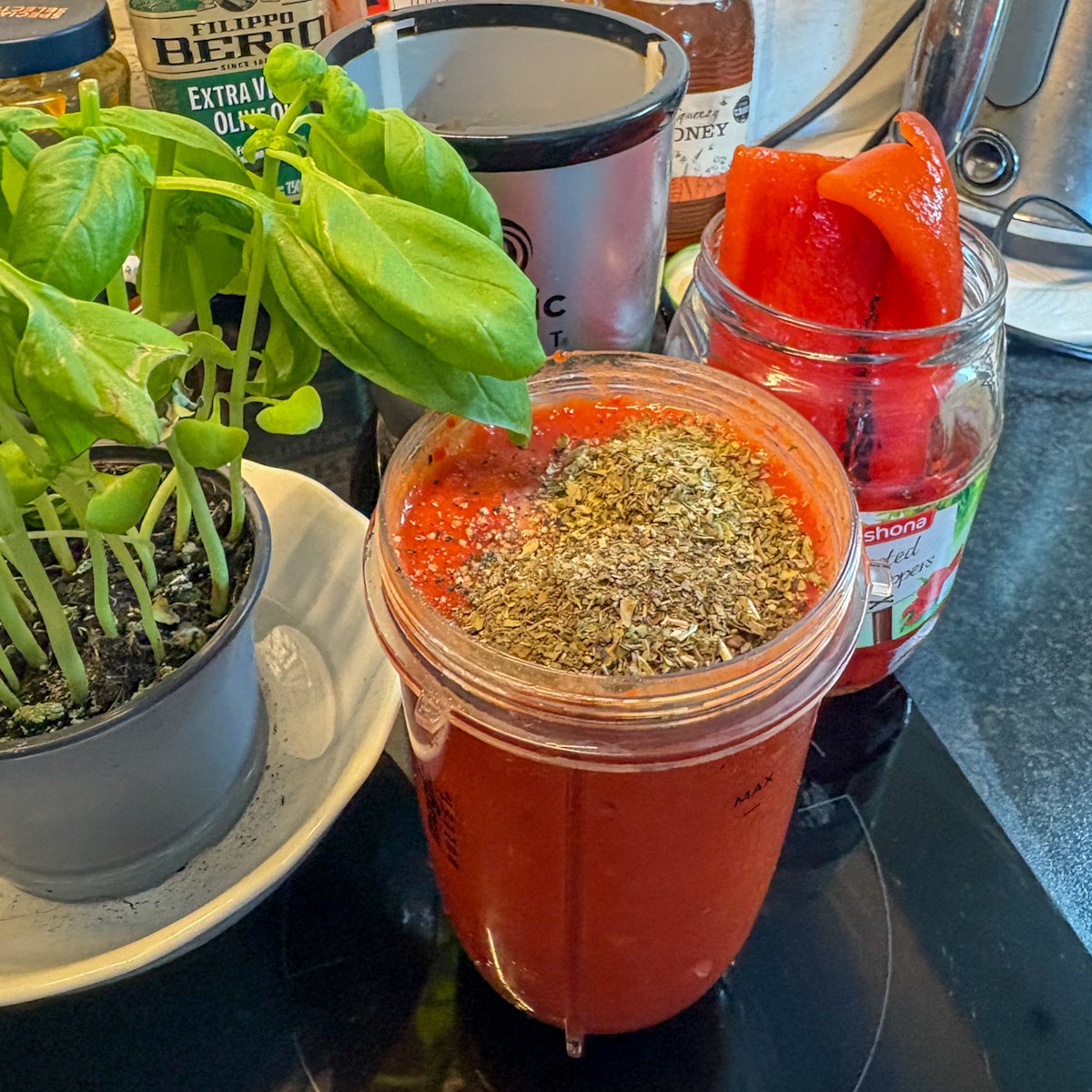 Blended roasted pepper and tomato sauce topped with dried herbs, a basil plant and peppers beside it