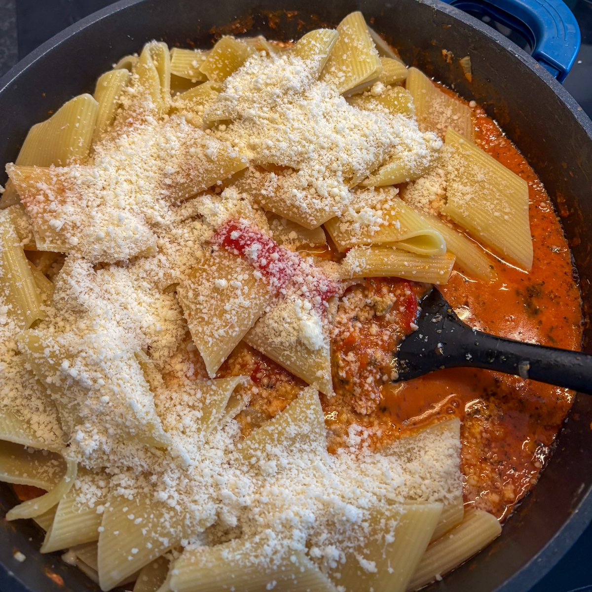 Rigatoni and grated parmesan added to roasted pepper sauce in a large black pan