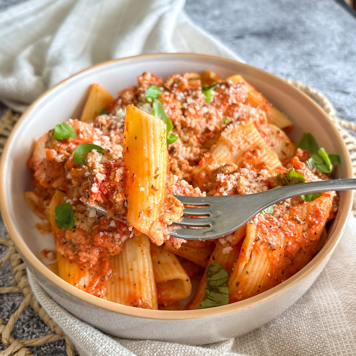 Fork lifting a rigatoni tube from a bowl of creamy turkey and roasted pepper pasta with basil