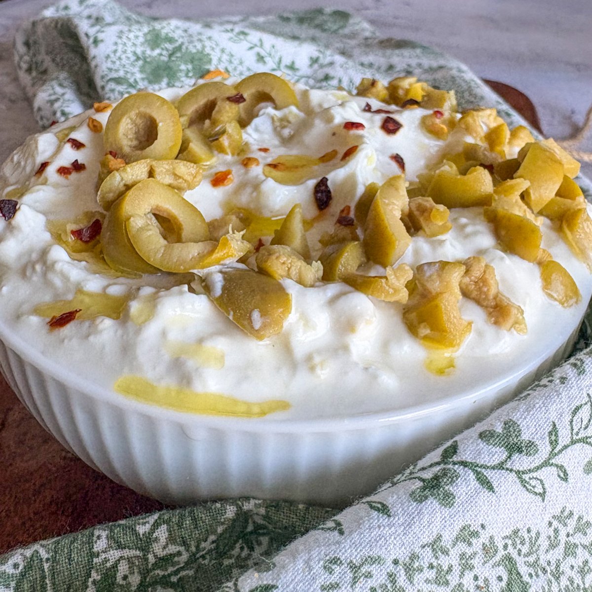 Easy whipped feta dip topped with sliced green olives, red chilli flakes and an olive oil drizzle, photographed from above with seasoned pitta chips on a green patterned cloth.