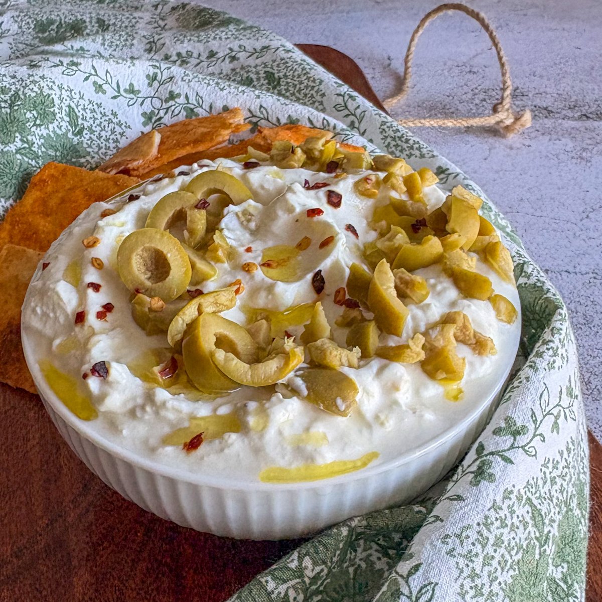 Creamy whipped feta dip in a ribbed white bowl with a pitta chip standing in the thick dip, scattered green olives and chilli flakes on top, and toasted pitta chips behind on a wooden board.