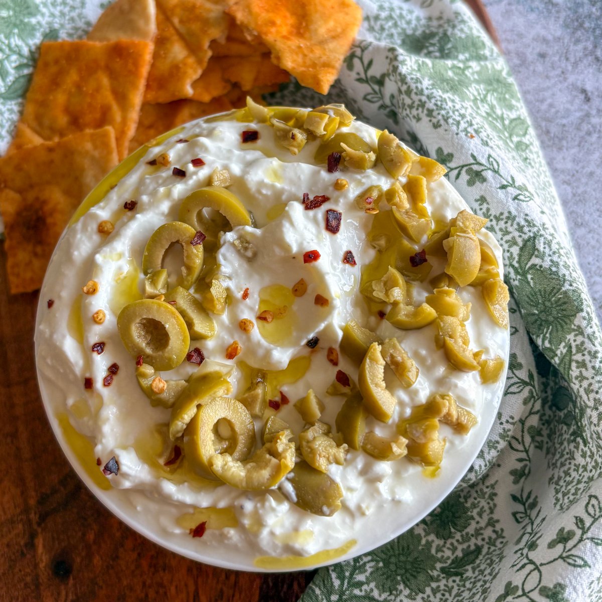 Top-down view of lemon whipped feta dip with deep spoon swirls, sliced green olives, red pepper flakes and glossy olive oil, served with toasted pitta chips on a rustic wooden board.