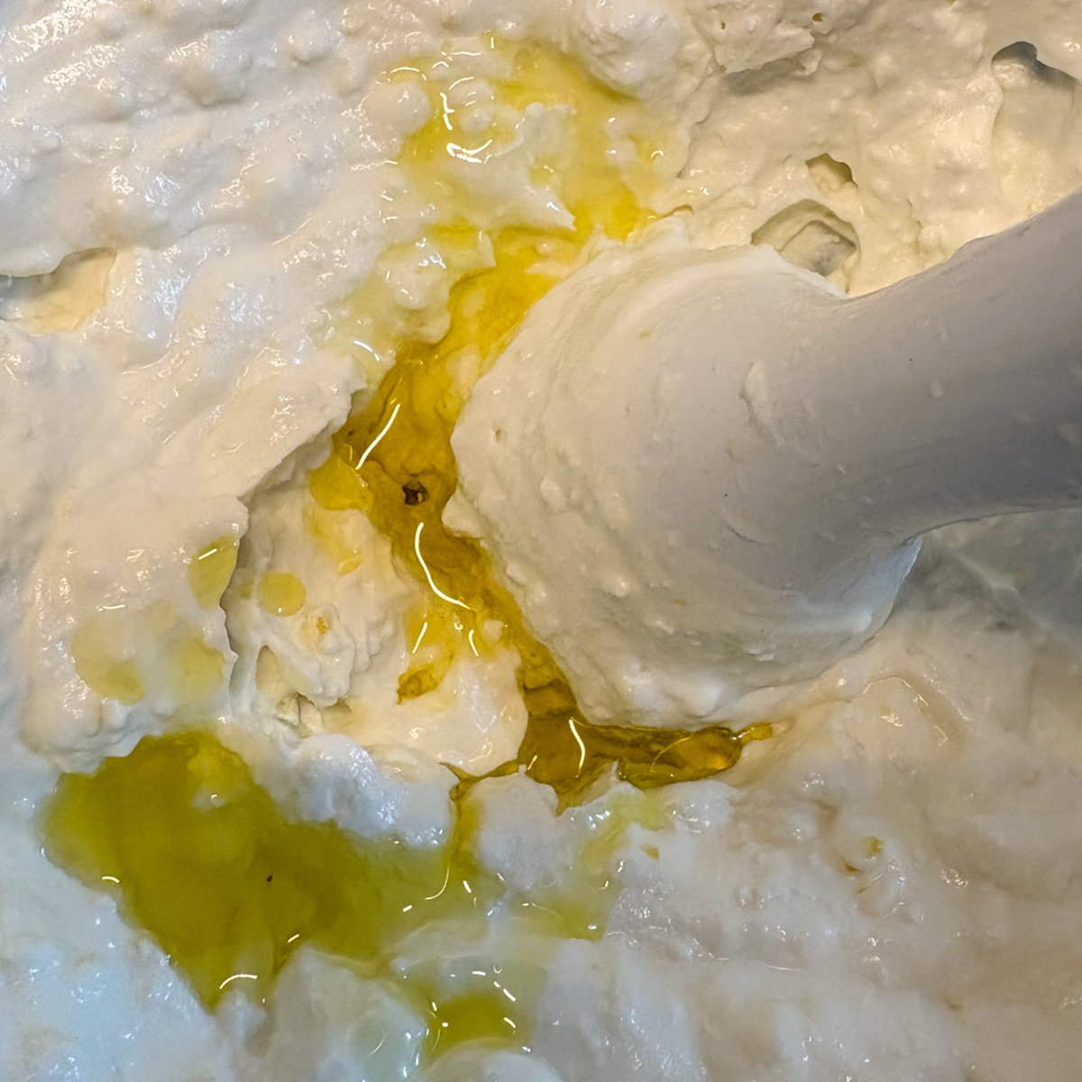Extra-virgin olive oil being blended into whipped feta with a hand blender, the oil pooling golden against the thick, creamy white mixture in the bowl.