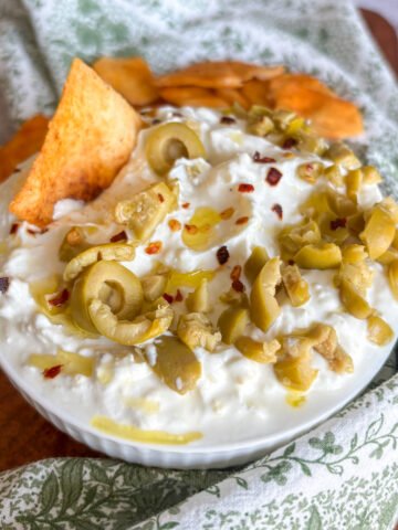 Whipped feta dip topped with sliced green olives, red chilli flakes and olive oil, with a golden pitta chip tucked into the creamy surface and more crisp pitta chips beside the bowl on a green floral cloth.