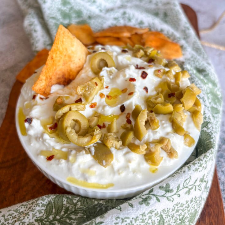 Whipped feta dip topped with sliced green olives, red chilli flakes and olive oil, with a golden pitta chip tucked into the creamy surface and more crisp pitta chips beside the bowl on a green floral cloth.