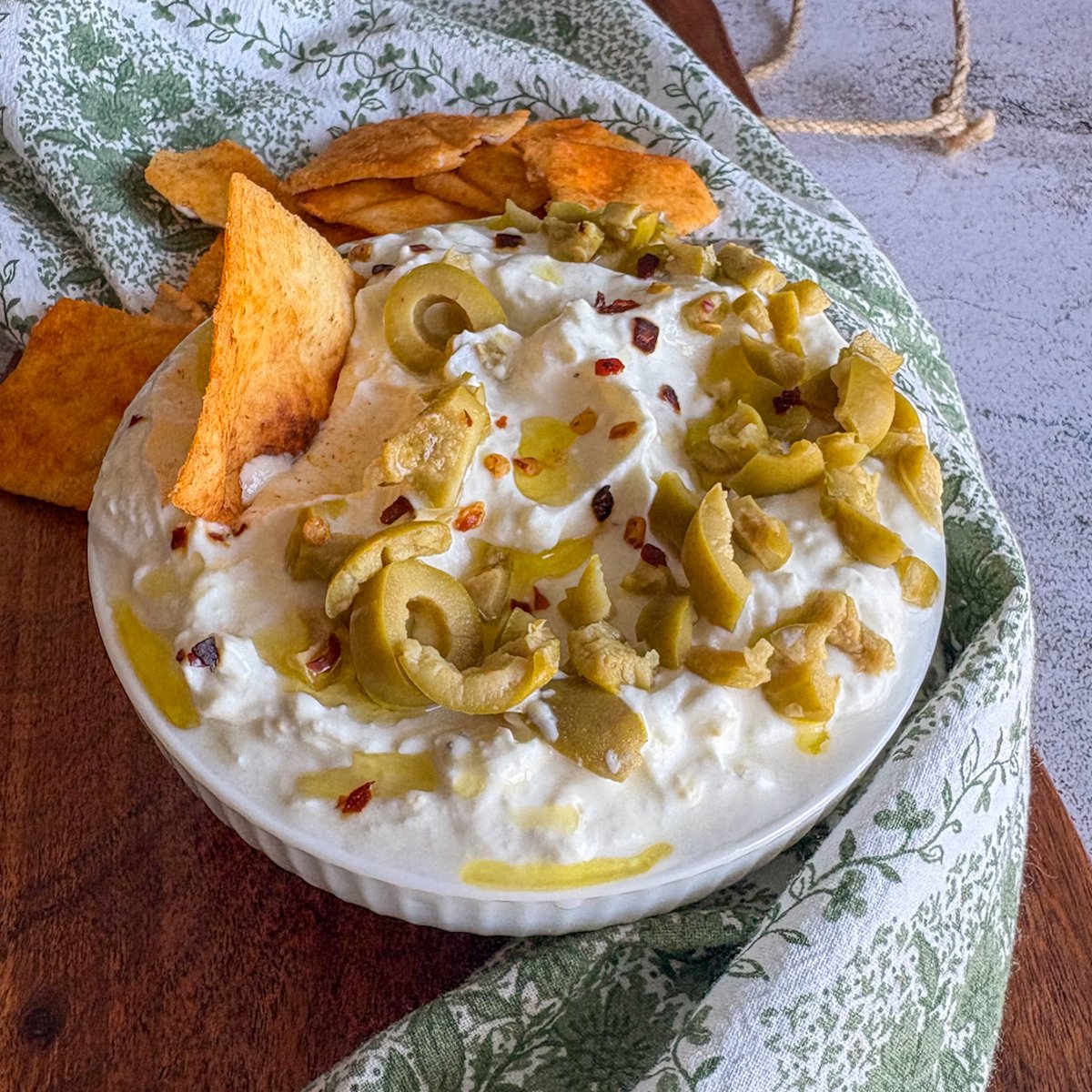Overhead view of whipped feta dip swirled in a white bowl, finished with crushed green olives, pools of olive oil and red pepper flakes, with a pile of seasoned pitta chips at the top of the frame.