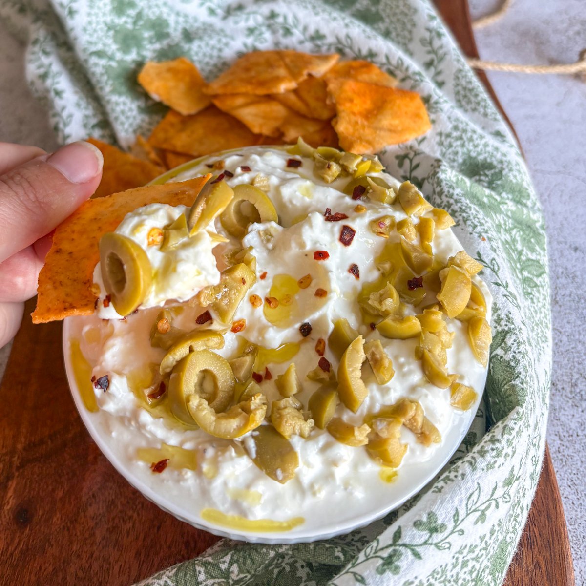 Hand scooping whipped feta dip with a crisp pitta chip, an olive slice riding on the creamy scoop, over a bowl topped with green olives, chilli flakes and golden olive oil.