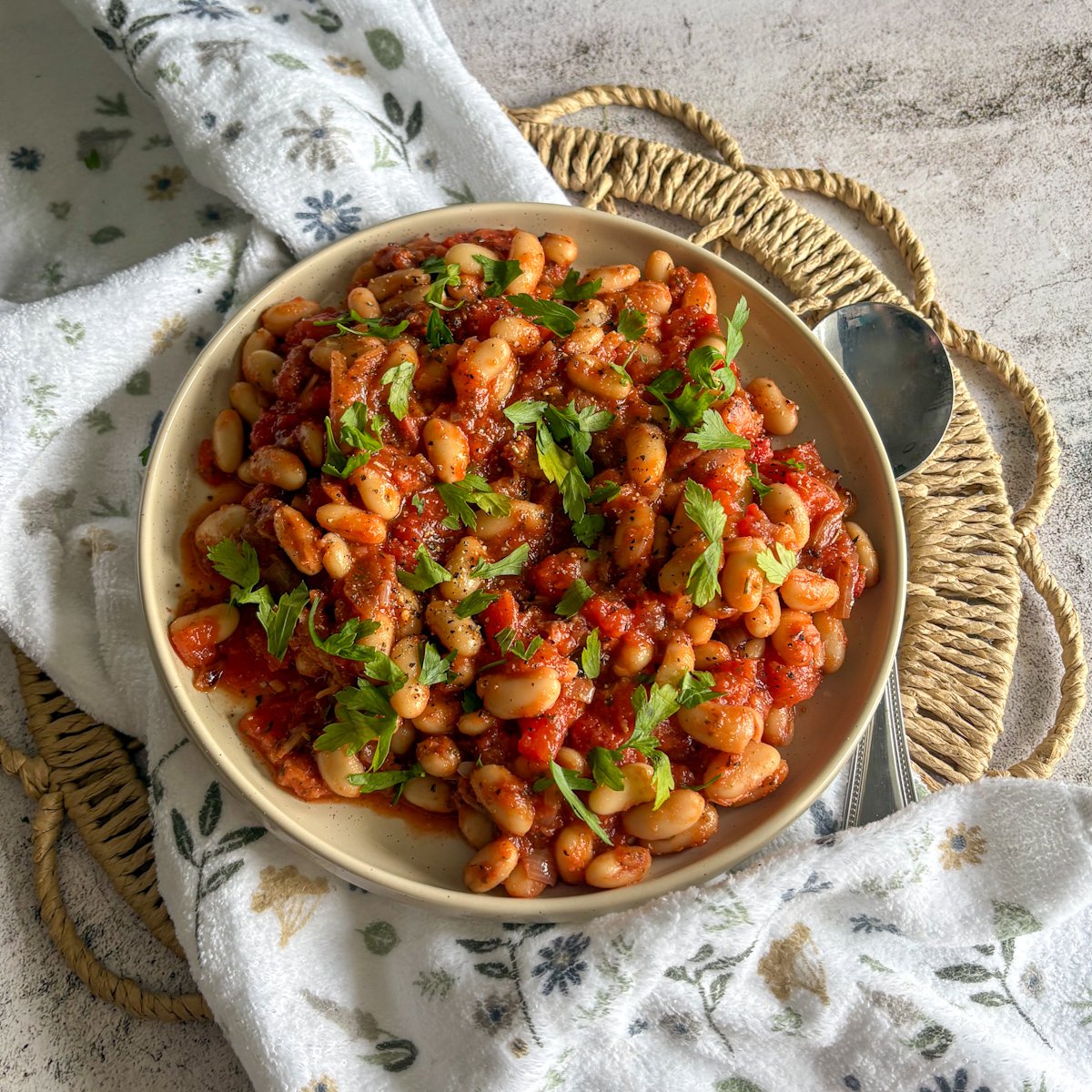 White beans and bacon in tomato sauce with a spoon on a woven mat and floral napkin
