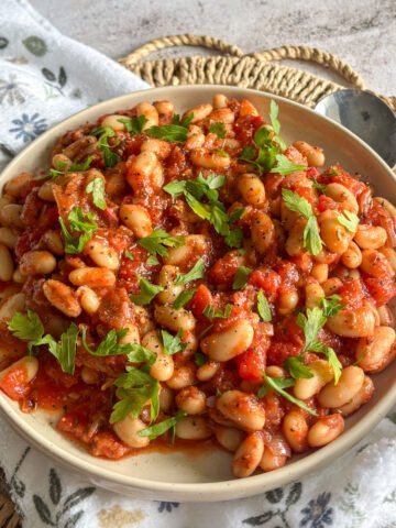 Bowl of white beans and bacon in tomato sauce on a woven mat with a floral cloth