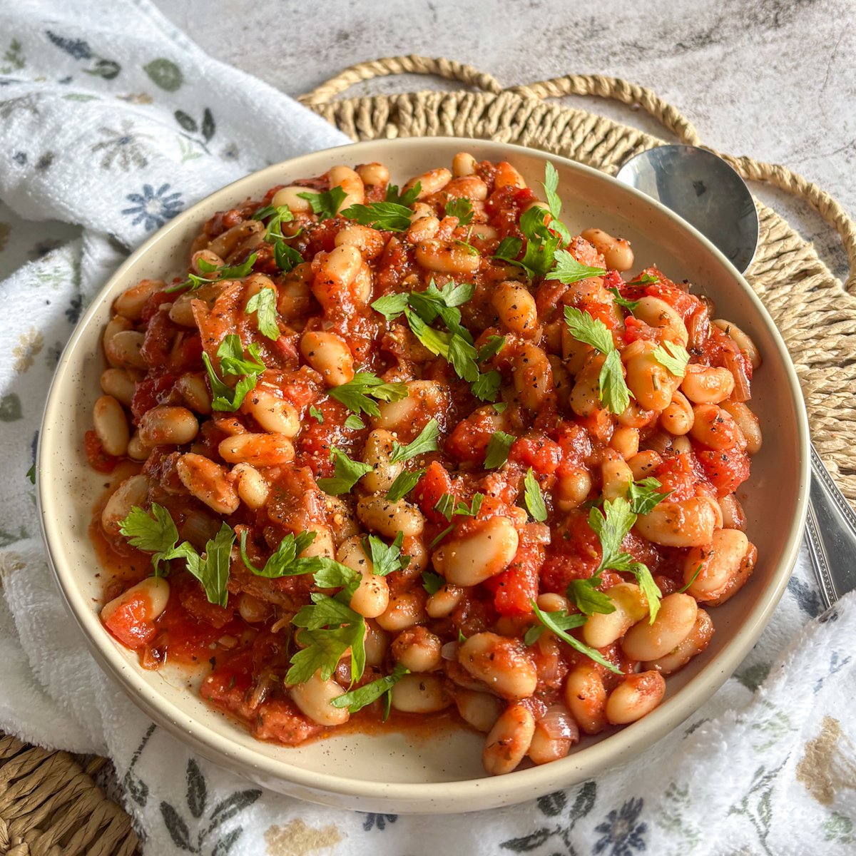 Bowl of white beans and bacon in tomato sauce on a woven mat with a floral cloth