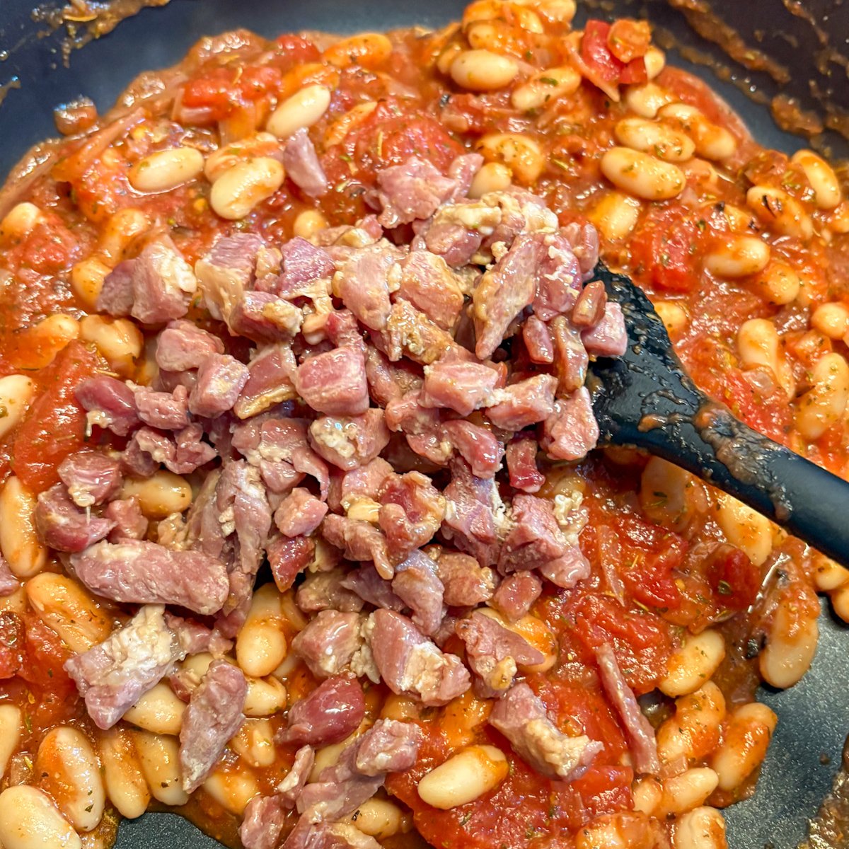 Chopped bacon stirred into white beans and tomato sauce in a skillet with a spatula