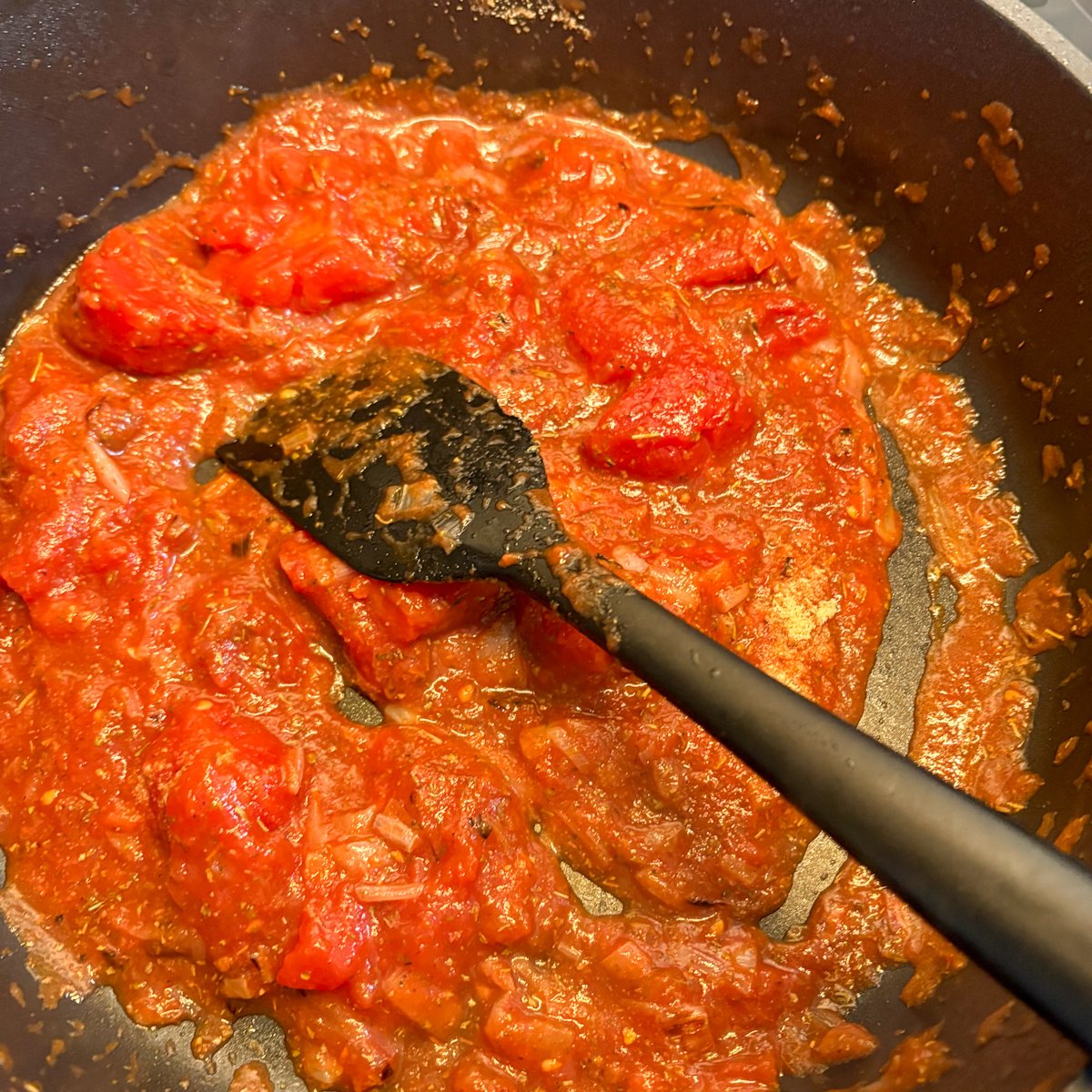 Crushed plum tomatoes cooking down in a skillet with a spatula for white beans and bacon