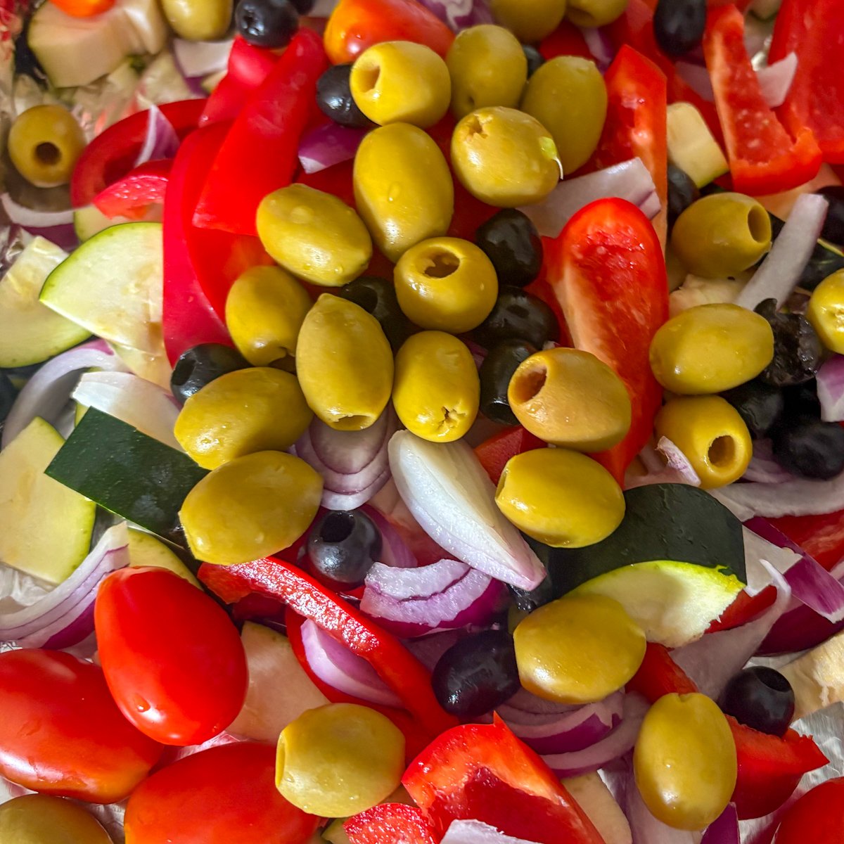 Vegetables for baked Mediterranean meatballs on a foil-lined tray before seasoning, including large pitted green olives, black olives, sliced red peppers, red onion, courgette pieces and mini plum tomatoes.