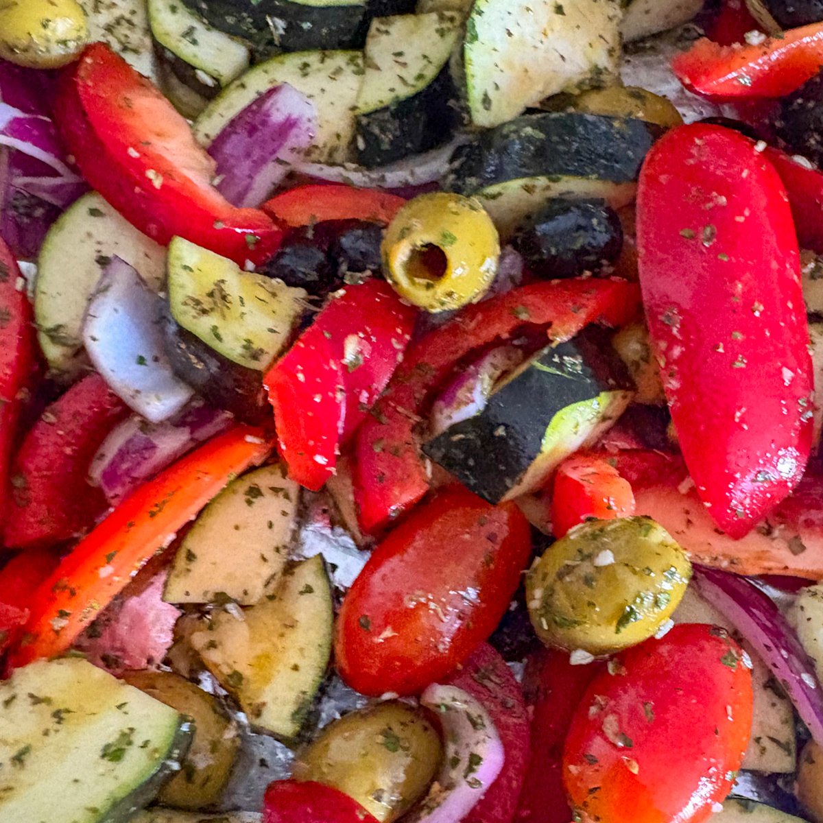 Mediterranean meatballs with roasted vegetables preparation, showing raw courgette quarters, red pepper wedges, sliced red onion, mini plum tomatoes and green and black olives glossy with olive oil and sprinkled with dried herbs before roasting.