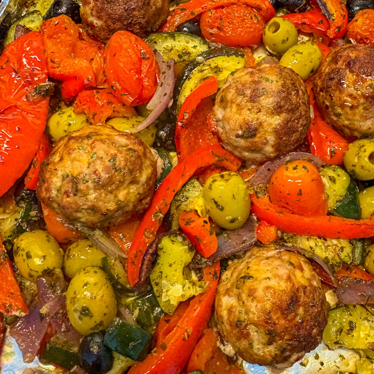 Mediterranean meatball traybake straight from the oven on a foil-lined baking tray, with golden meatballs among blistered mini plum tomatoes, roasted red peppers, courgettes, red onion and green and black olives coated in dried herbs.