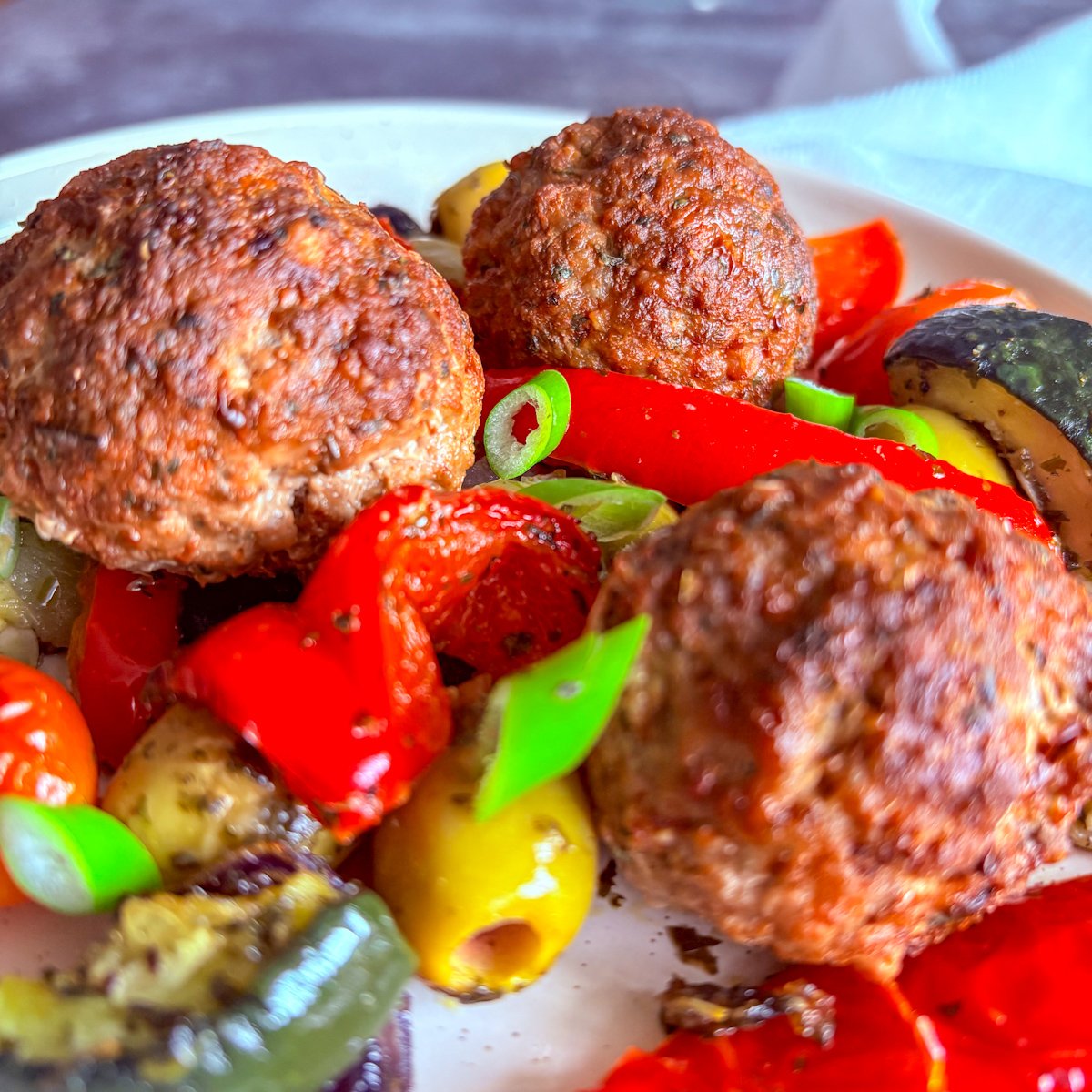 Healthy baked meatballs recipe overhead shot, a cream bowl of Mediterranean meatballs with roasted red peppers, green and black olives, blistered mini plum tomatoes and courgette on a woven placemat with a striped tea towel.