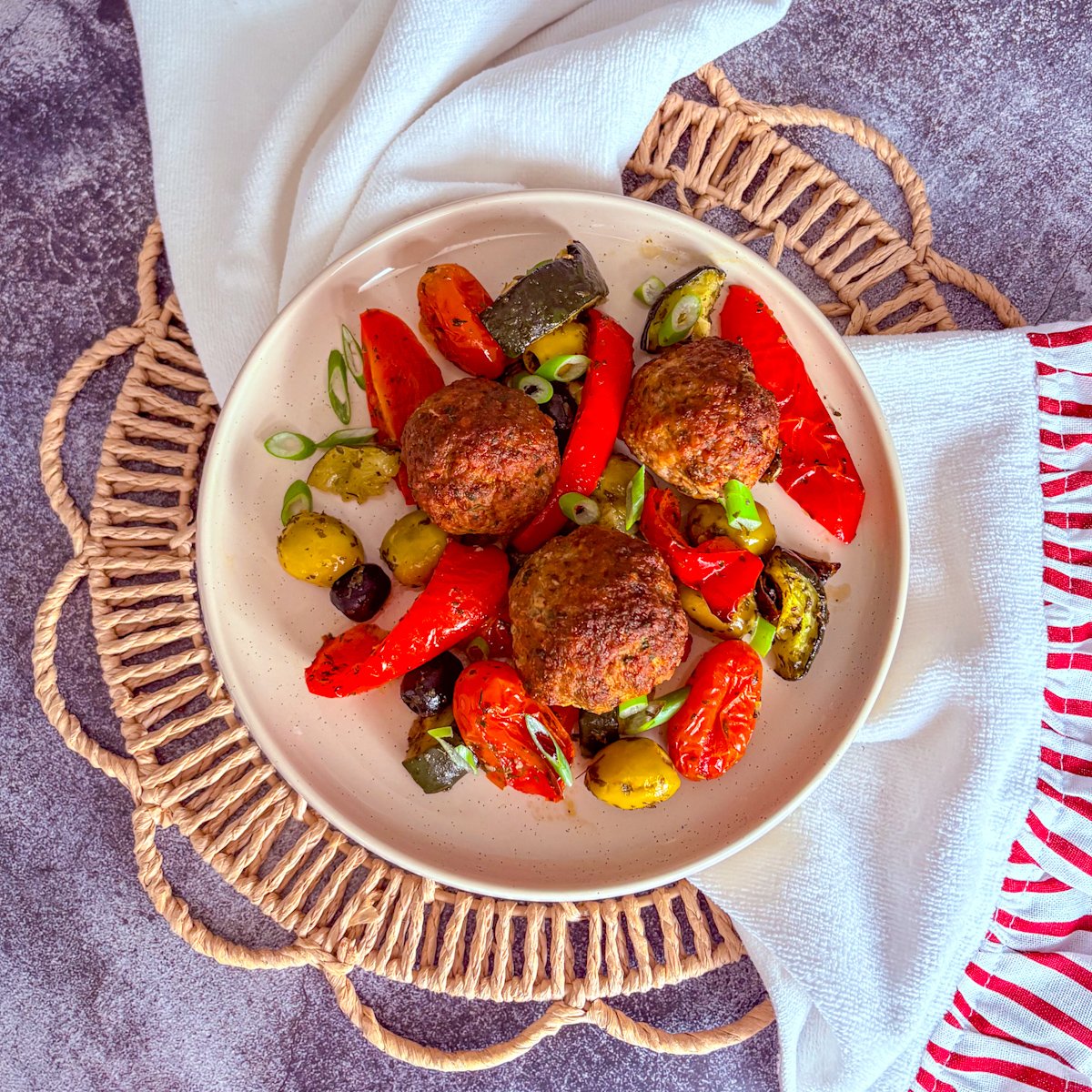 Baked Mediterranean meatballs overhead close-up in a cream bowl, with three browned meatballs surrounded by blistered mini plum tomatoes, roasted red peppers, soft olives, charred courgette and spring onion slices.